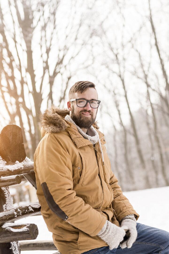 Ein Mann mit Brille und Winterjacke sitzt auf einem Holzzaun im verschneiten Wald.