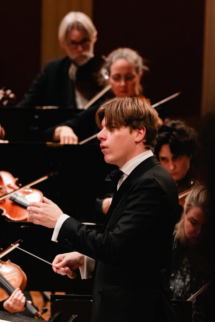 Ein Dirigent - Klaus Mäkelä -  leitet ein Orchester, dessen Musiker konzentriert Streichinstrumente spielen.