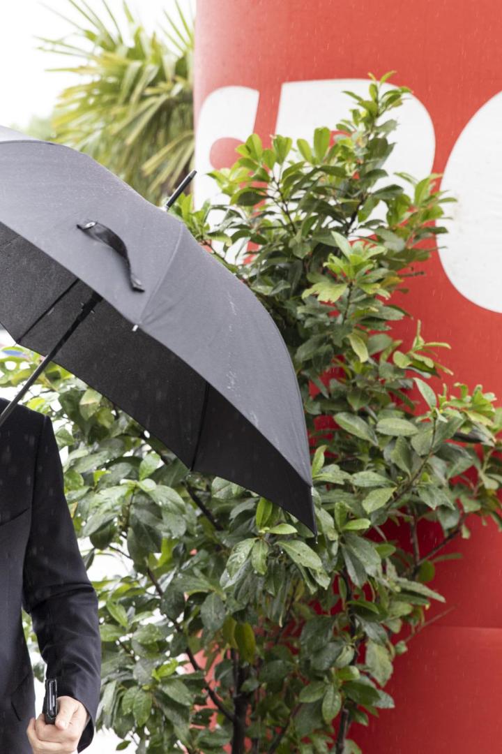 Ein Mann mit Anzug und Krawatte hält einen Regenschirm im Regen vor einem roten Hintergrund mit einem weißen "Ö".