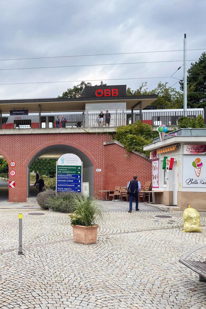Ein ÖBB-Zug fährt über eine rote Backsteinbrücke mit Durchgängen, neben einem Platz mit einem Eisstand namens "Bella Rosa".
