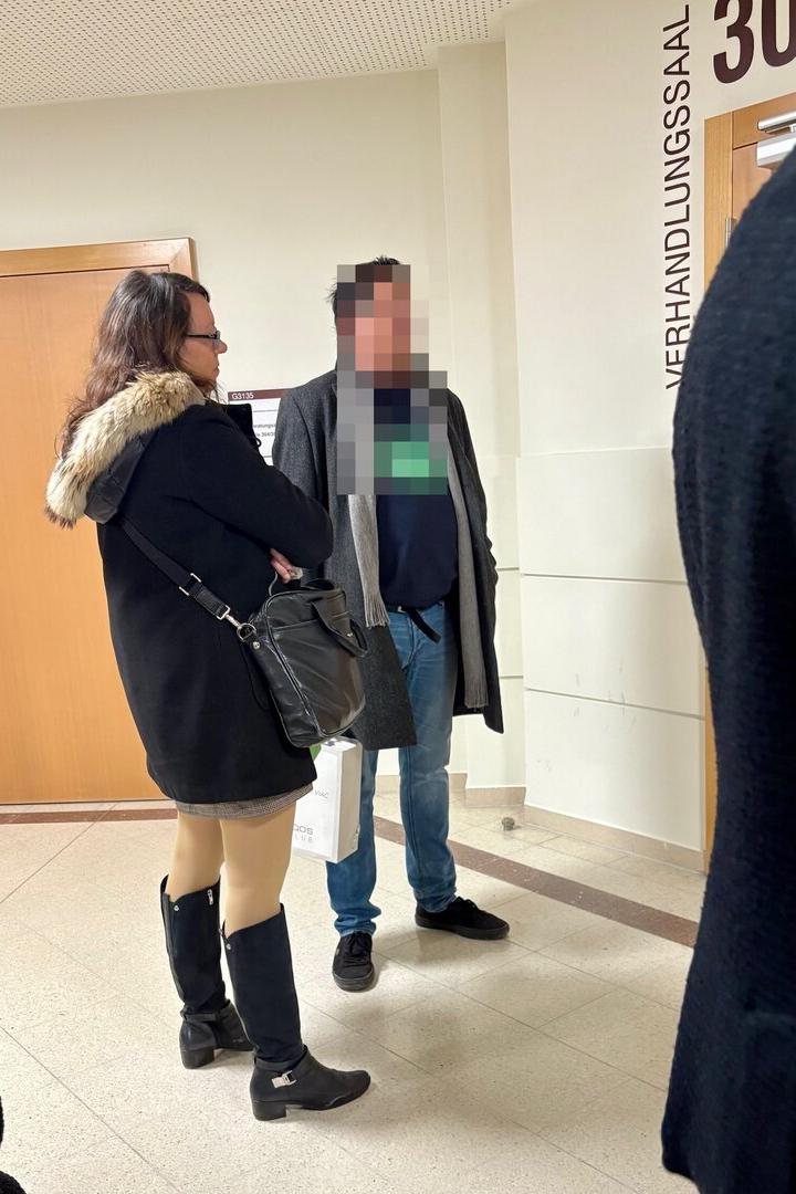 Zwei Personen stehen in einem Flur vor einer Tür mit der Aufschrift „Verhandlungssaal“.