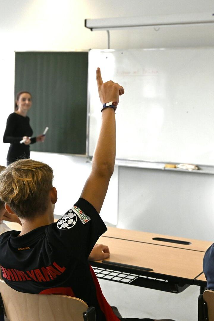 Mehrere Schüler sitzen im Klassenraum und melden sich, während eine Lehrerin vorne an der Tafel steht.
