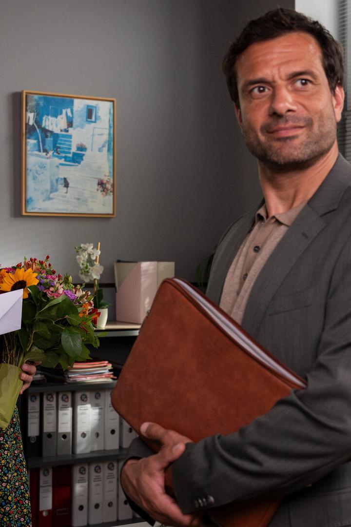 Eine Frau (Marisa Burger/ Frau Stockl) hält einen Blumenstrauß und einen Umschlag, ein Mann im Anzug steht mit einer Ledermappe daneben.