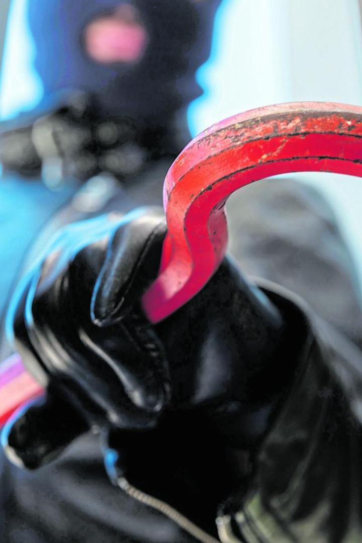 Eine vermummte Person mit Handschuhen hebelt mit einem roten Brecheisen ein Fenster auf.