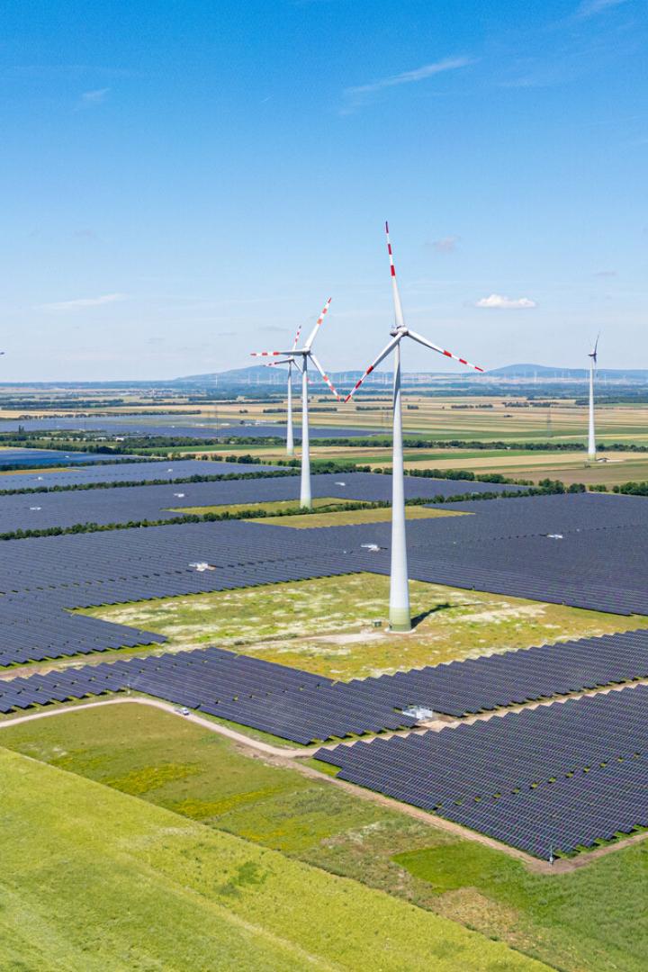 Mehrere Windräder und große Solarfelder stehen auf einer weitläufigen grünen Landschaft unter blauem Himmel.