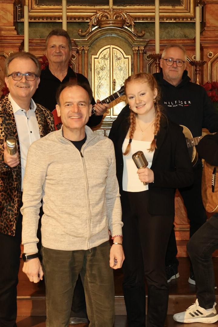 Eine Musikgruppe mit Gitarren und Mikrofonen posiert vor einem verzierten Altar und Weihnachtsbäumen.