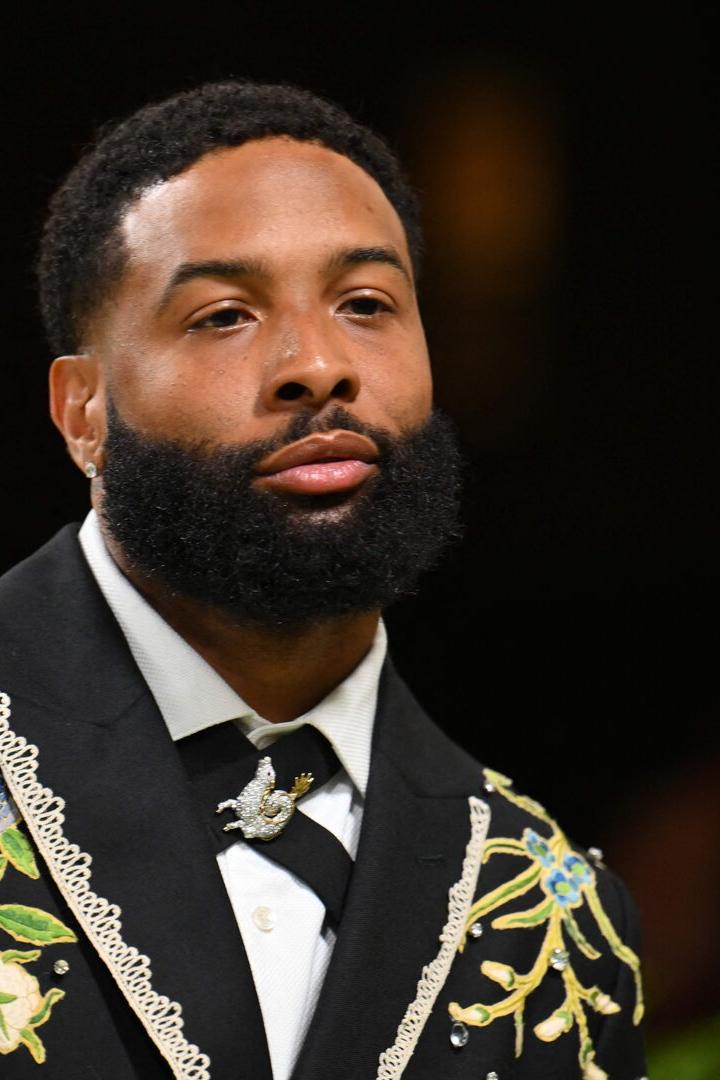 Ein Mann mit Vollbart trägt einen schwarzen Anzug mit floralen Stickereien und Schmuckdetails.