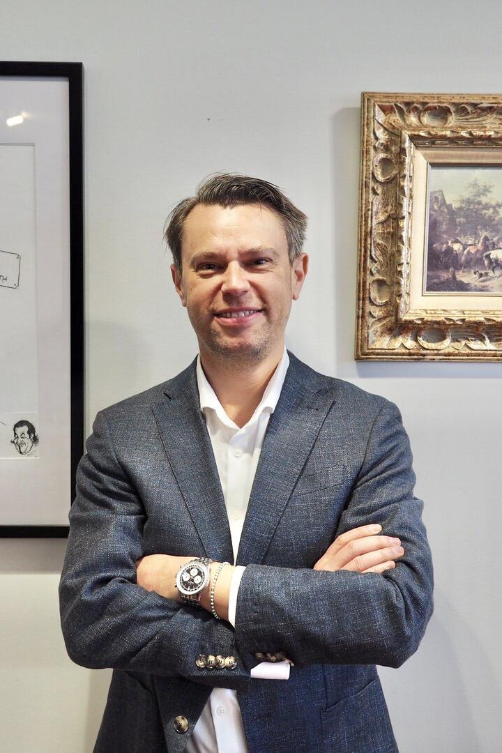 Ein Mann im Anzug steht lächelnd mit verschränkten Armen vor zwei gerahmten Bildern an einer Wand.