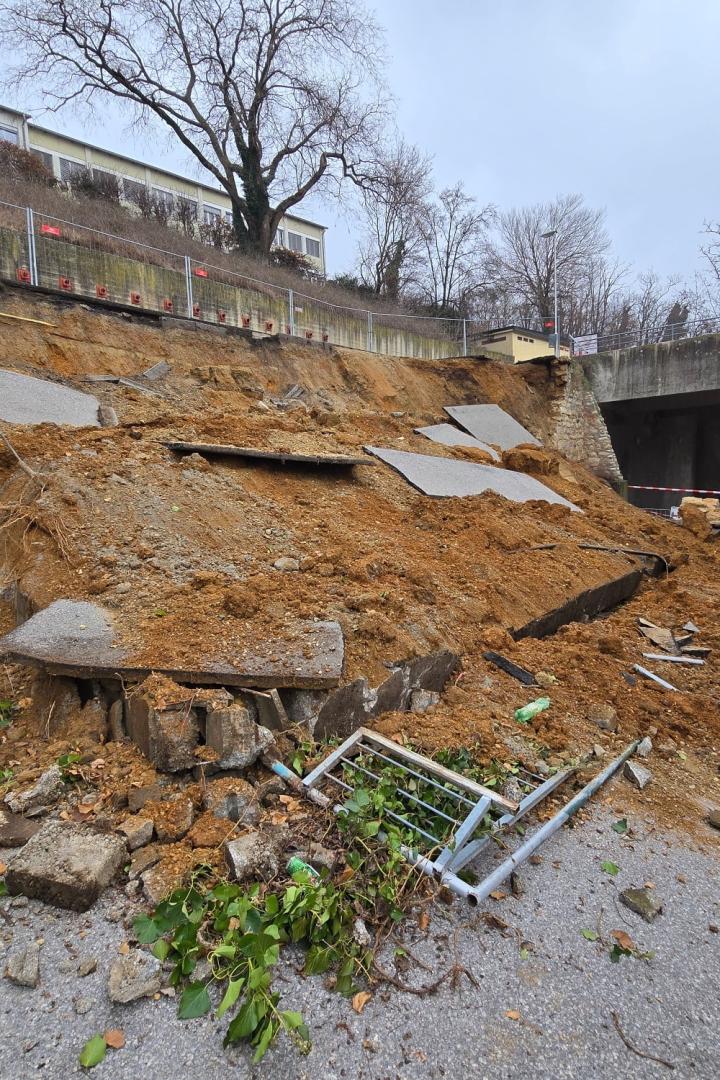 Eingestürzte Stützmauer mit Erdrutsch und Absperrungen im Bereich eines Wohngebäudes.