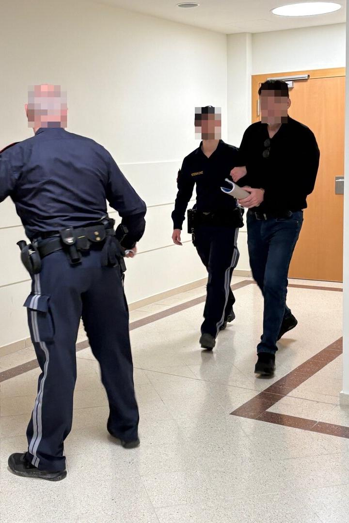 Zwei Polizisten führen einen Mann mit gefesselten Händen durch einen Flur zu einer Tür.