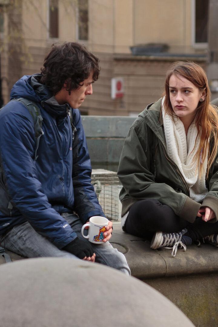 Zwei junge Menschen sitzen auf einer steinernen Umrandung, einer hält eine Tasse, beide wirken nachdenklich.