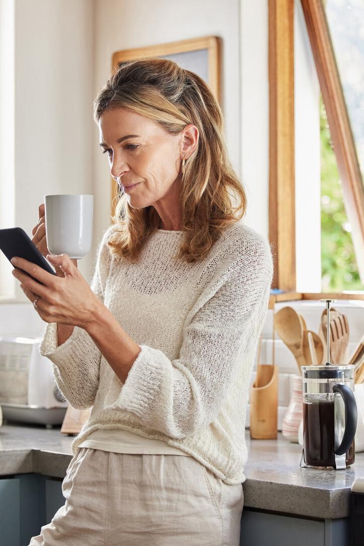 Coffee, phone and senior woman in kitchen browsing social media, text message or online app at home. Relax, tea and reading news or mobile online web surfing with female from Canada on 5g smartphone