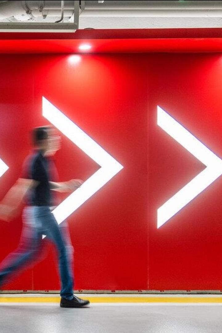 Eine Person geht vor einer roten Wand mit vier großen, leuchtenden Pfeilen nach rechts entlang.