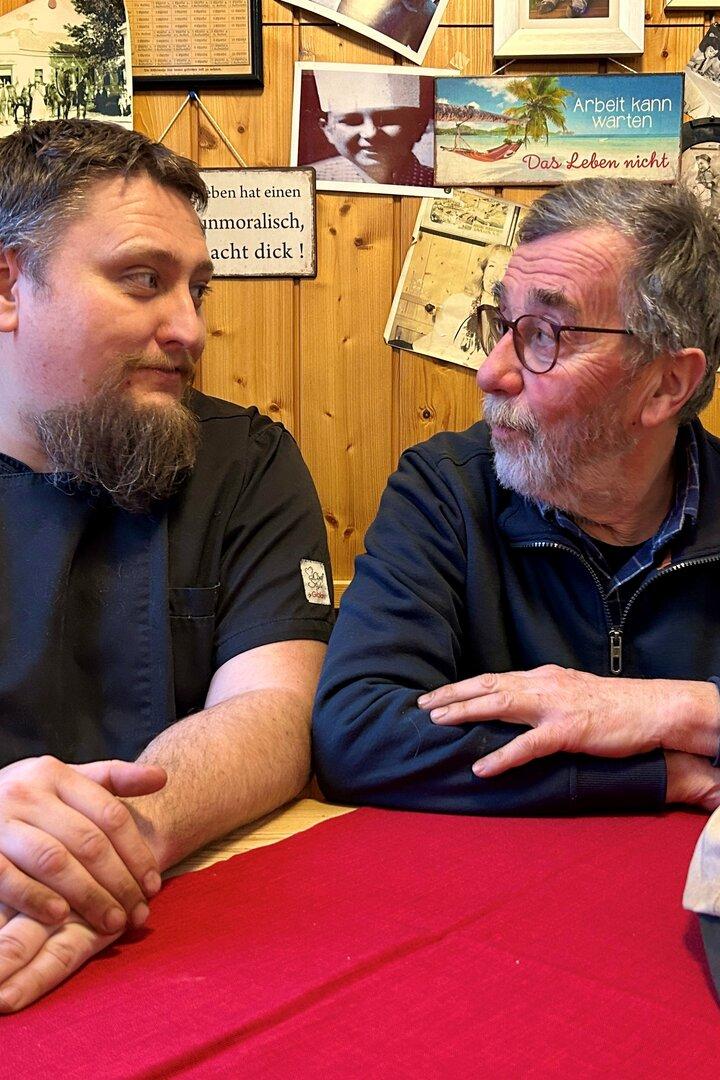 Zwei Männer sitzen an einem Tisch mit roter Decke, im Hintergrund Holzvertäfelung mit Dekoration.