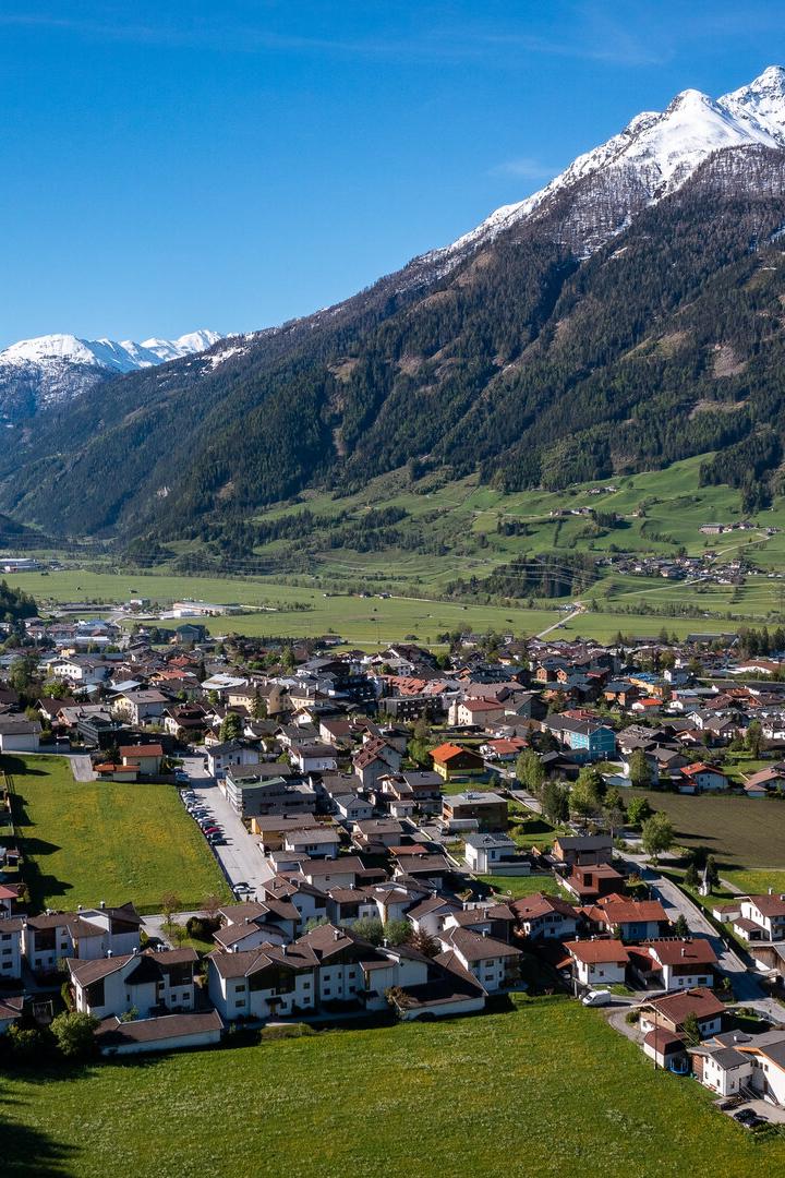 Ein Dorf liegt in einem Tal, umgeben von grünen Hängen und schneebedeckten Bergen.