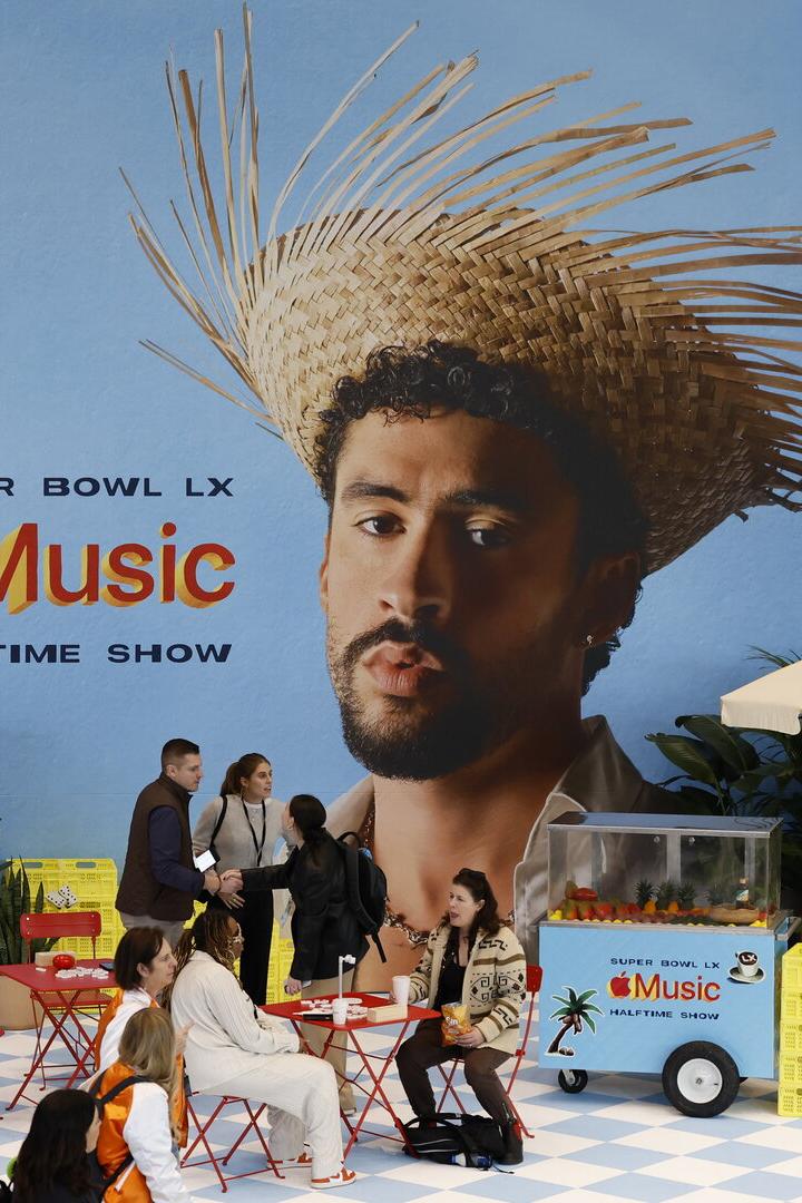 Mehrere Menschen sitzen an roten Tischen vor einer großen Werbewand für den Super Bowl LX Apple Music Halftime Show.