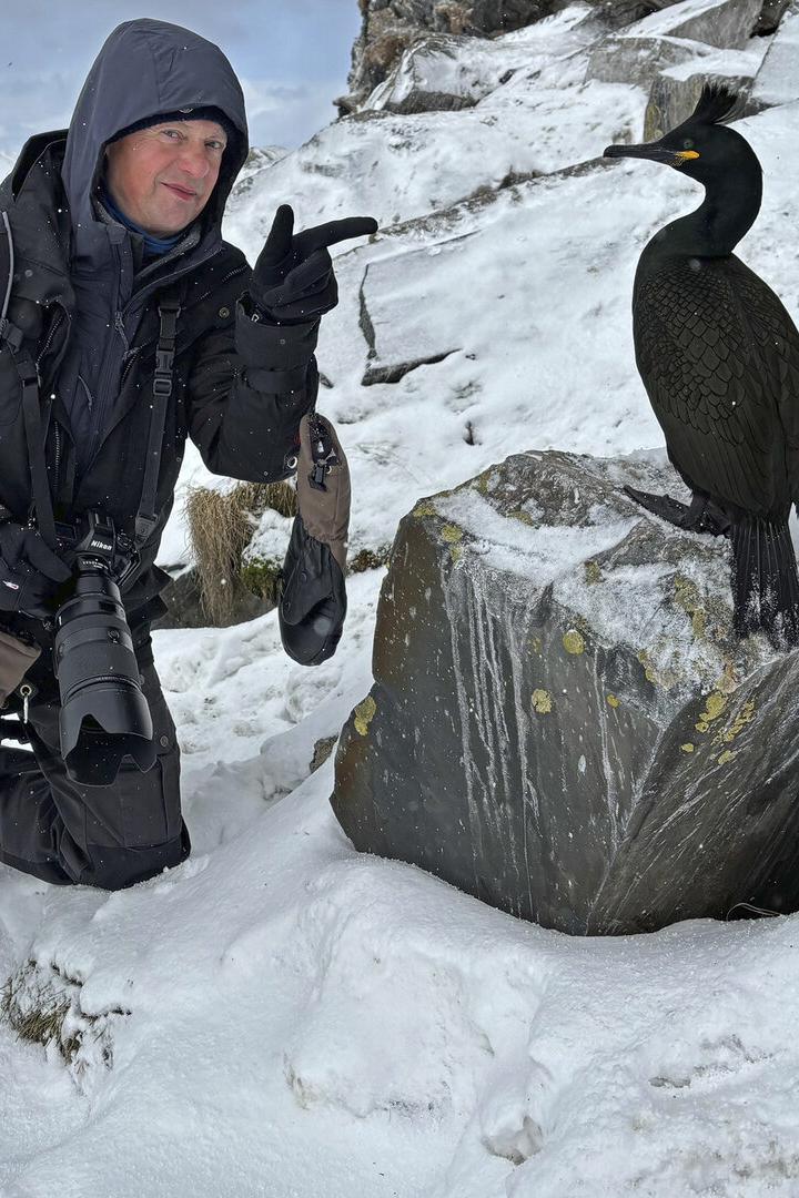 Ein Mann mit Kamera kniet im Schnee neben einem Kormoran auf einem Felsen.