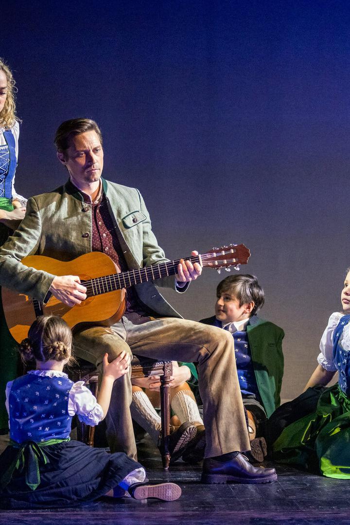 Ein Mann spielt Gitarre, umgeben von Kindern und einer Frau in traditionellen Trachten auf einer Bühne.