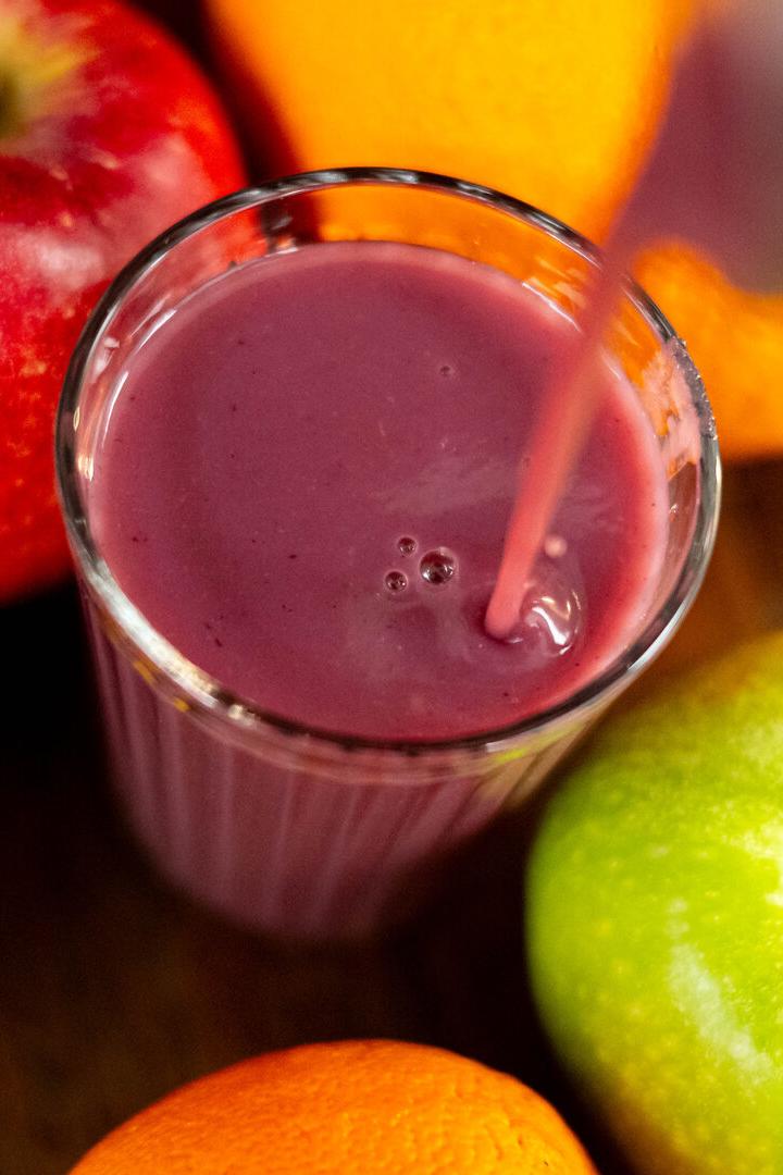 Ein Glas lila Smoothie wird umgeben von Apfel, Orange und Zitrone eingeschenkt.