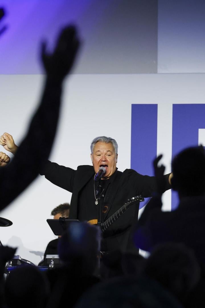 Ein Sänger mit Gitarre steht vor Publikum mit erhobenen Händen auf einer Bühne.