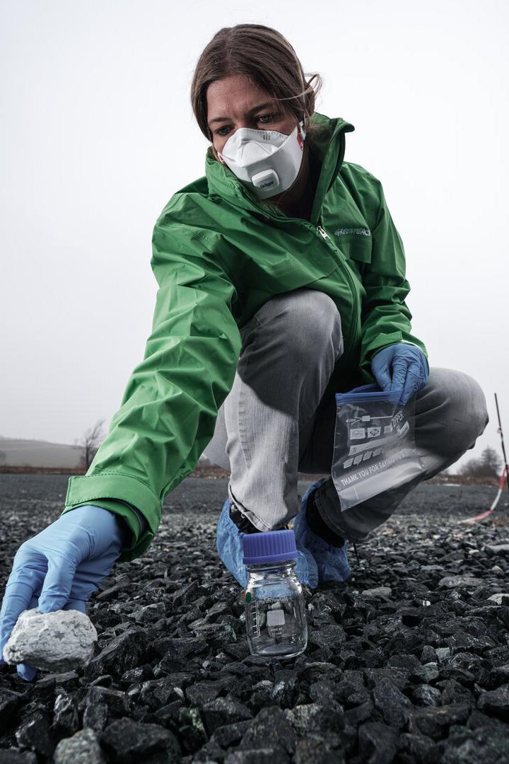 Eine Frau mit Maske und Handschuhen sammelt eine Gesteinsprobe in einem Glasgefäß.