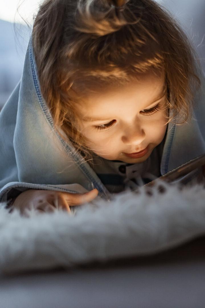 Ein kleines Mädchen liegt in eine Decke gehüllt und schaut auf ein leuchtendes Handy.
