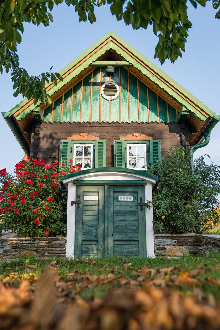 Ein kleines, dunkles Holzhaus mit grünen Akzenten und roten Rosenbüschen davor.