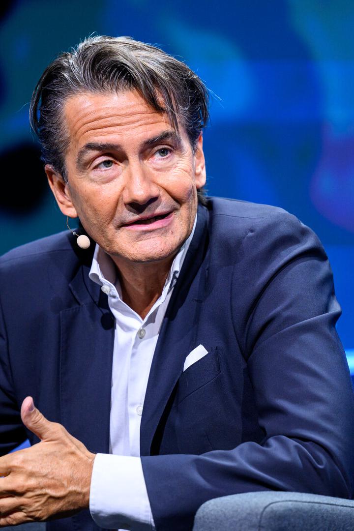 Ein Mann mit Mikrofon und dunklem Sakko gestikuliert vor blauem Hintergrund.