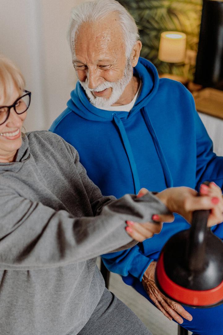 Ein älteres Paar trainiert gemeinsam mit Hantel und Kettlebell und lächelt dabei.