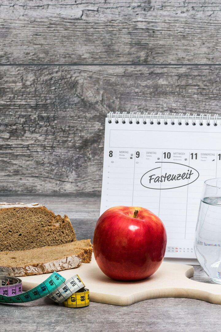 Ein Apfel, ein Glas Wasser, Brot, ein Maßband und ein Kalender mit dem Wort „Fastenzeit“ auf einem Holztisch.