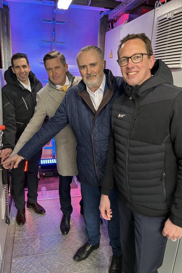 Vier Männer stehen in einem Raum mit technischer Ausrüstung und bedienen gemeinsam eine Steuerung.