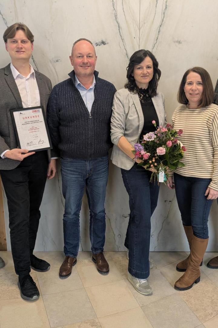 Sechs Personen stehen nebeneinander, eine Person hält eine Urkunde, eine andere einen Blumenstrauß.