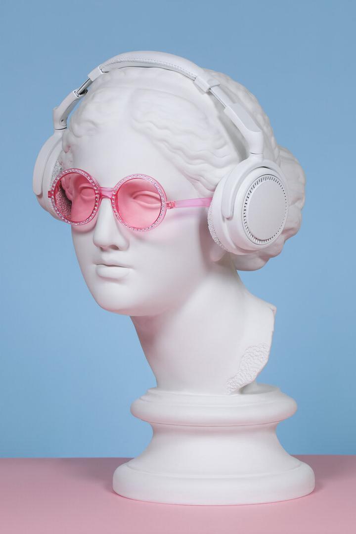 Eine weiße Büste trägt pinke Sonnenbrille und weiße Kopfhörer vor blauem Hintergrund auf rosa Fläche.