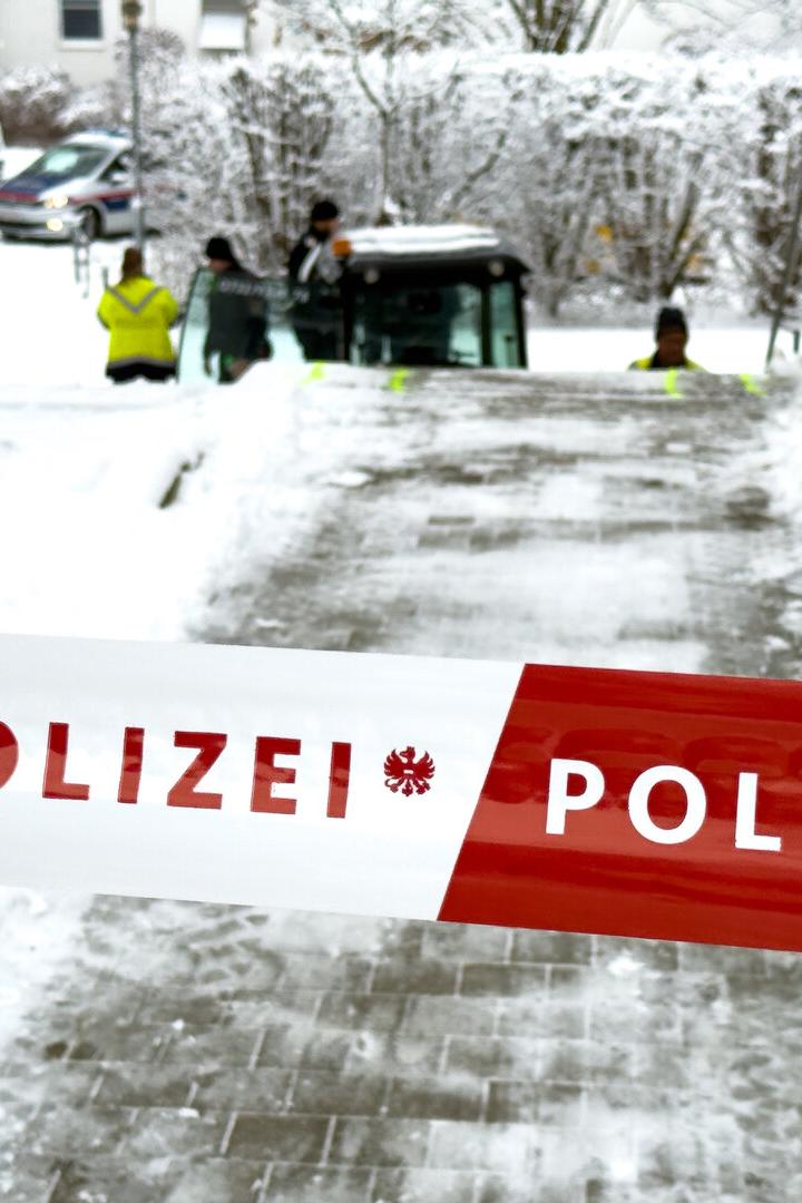 Ein rot-weißes Polizei-Absperrband versperrt einen verschneiten Weg, im Hintergrund sind Einsatzkräfte und ein Polizeiauto zu sehen.