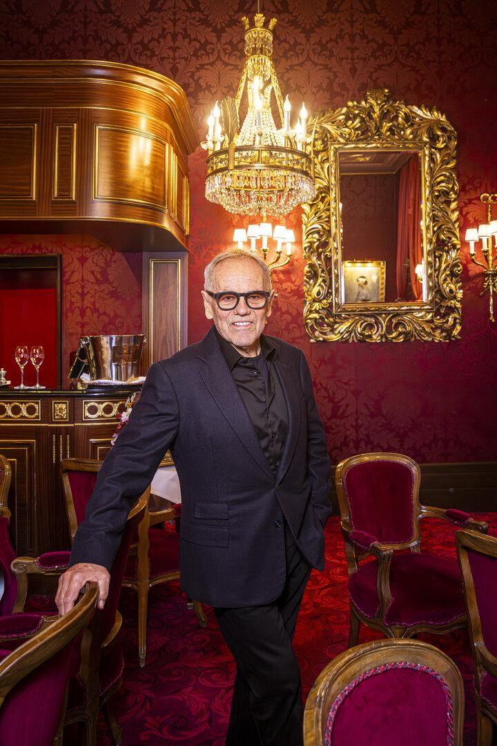 Wolfgang Puck mit Brille und dunklem Anzug steht in einem opulenten, rot dekorierten Raum mit goldenem Spiegel.
