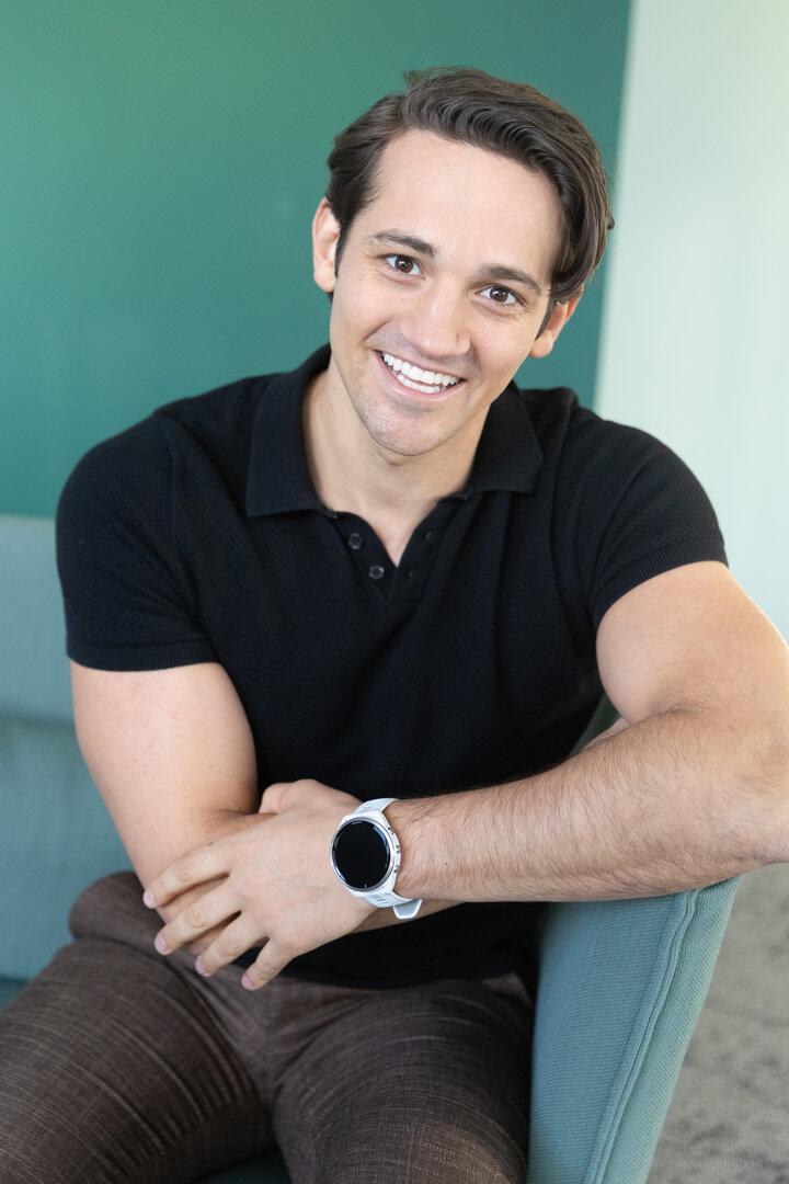 Ein junger Mann mit dunklem Haar und schwarzem Poloshirt sitzt lächelnd auf einem Stuhl und trägt eine weiße Armbanduhr.