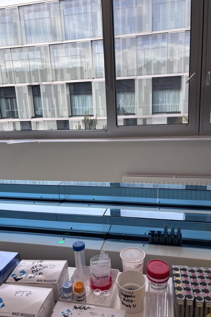 Laborutensilien und Probenröhrchen stehen auf einer Fensterbank, im Hintergrund ist ein modernes Gebäude zu sehen.