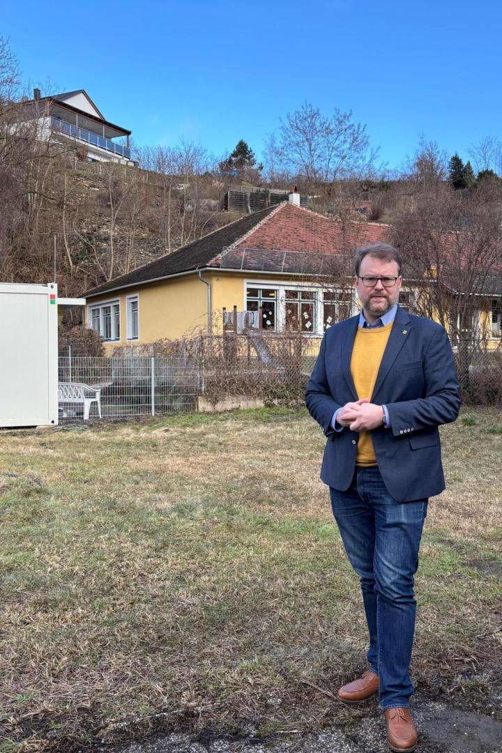 Florian Kamleitner steht vor dem Kindergarten und einer Containerklasse auf einer braun-grünen Wiese.