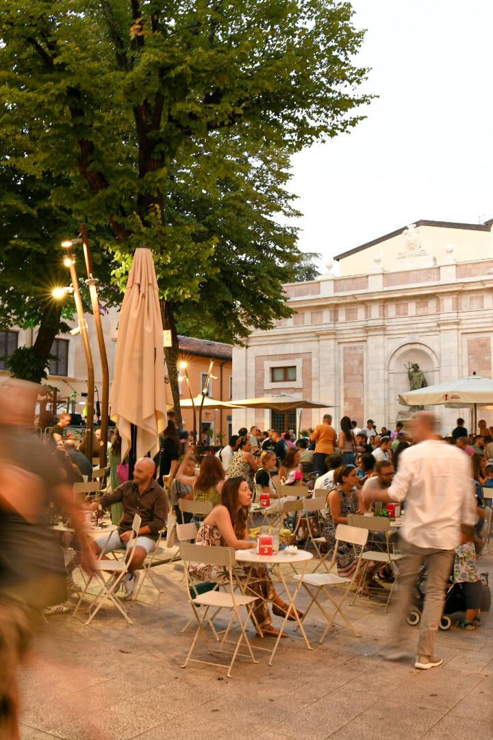Ein belebter Platz mit vielen Menschen an Tischen unter Sonnenschirmen vor einem Gebäude.