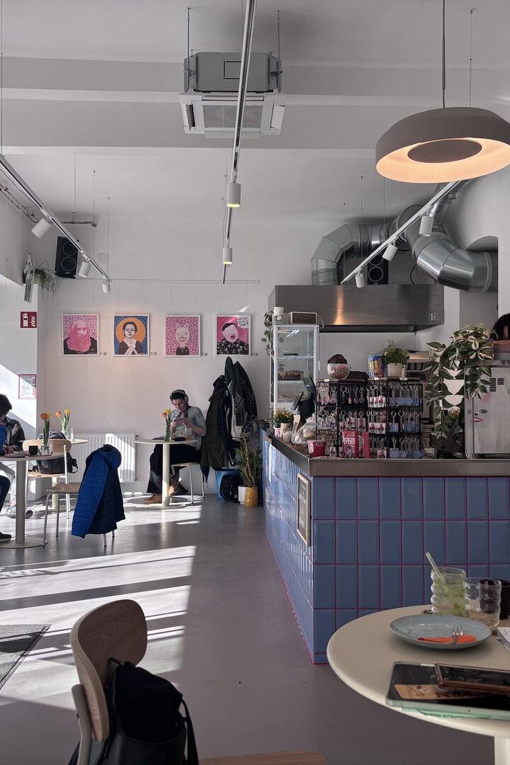 Helles Café mit wenigen Gästen, moderner Einrichtung, Blumen auf den Tischen und bunten Porträts an der Wand.