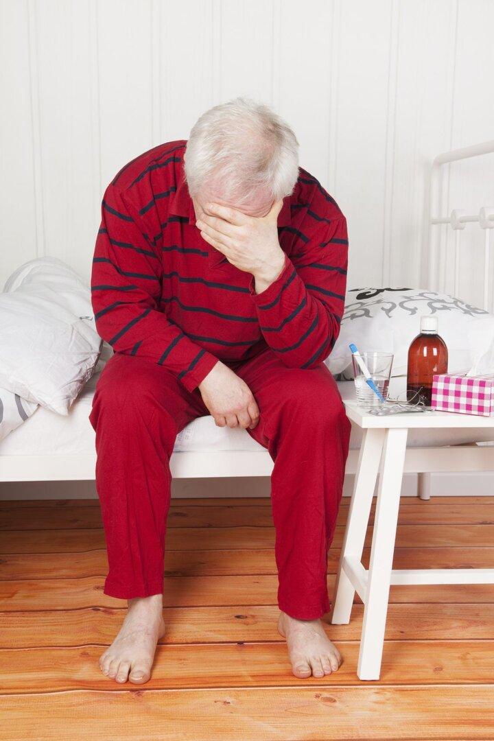 Älterer Mann im roten Schlafanzug sitzt erschöpft am Bettrand und hält den Kopf in der Hand, neben ihm Medikamente auf einem Tisch.