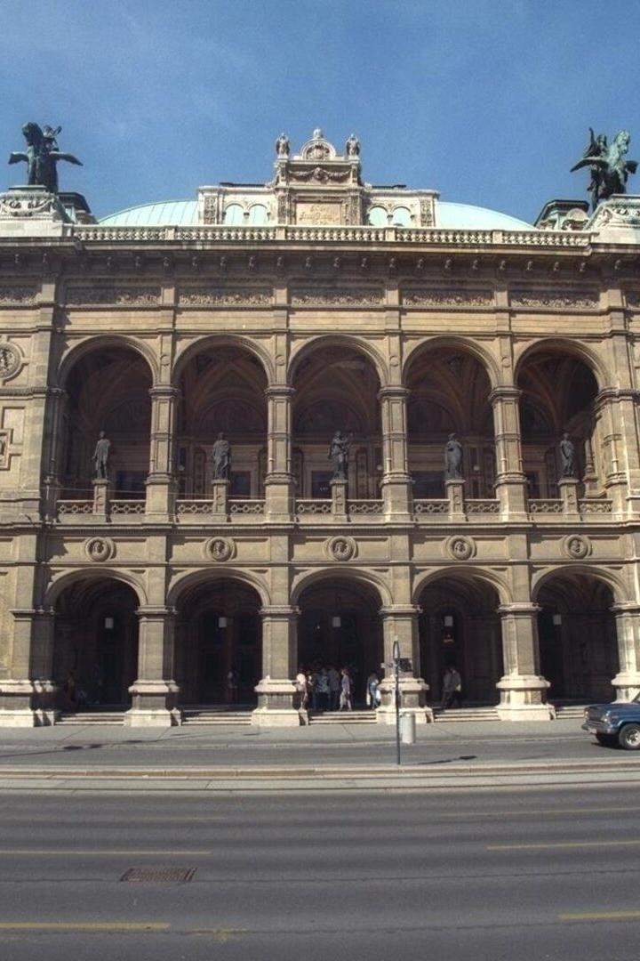 Prächtiges historisches Gebäude mit Arkaden, Statuen und Kuppel, davor fahren Autos und gehen Menschen entlang der Straße.