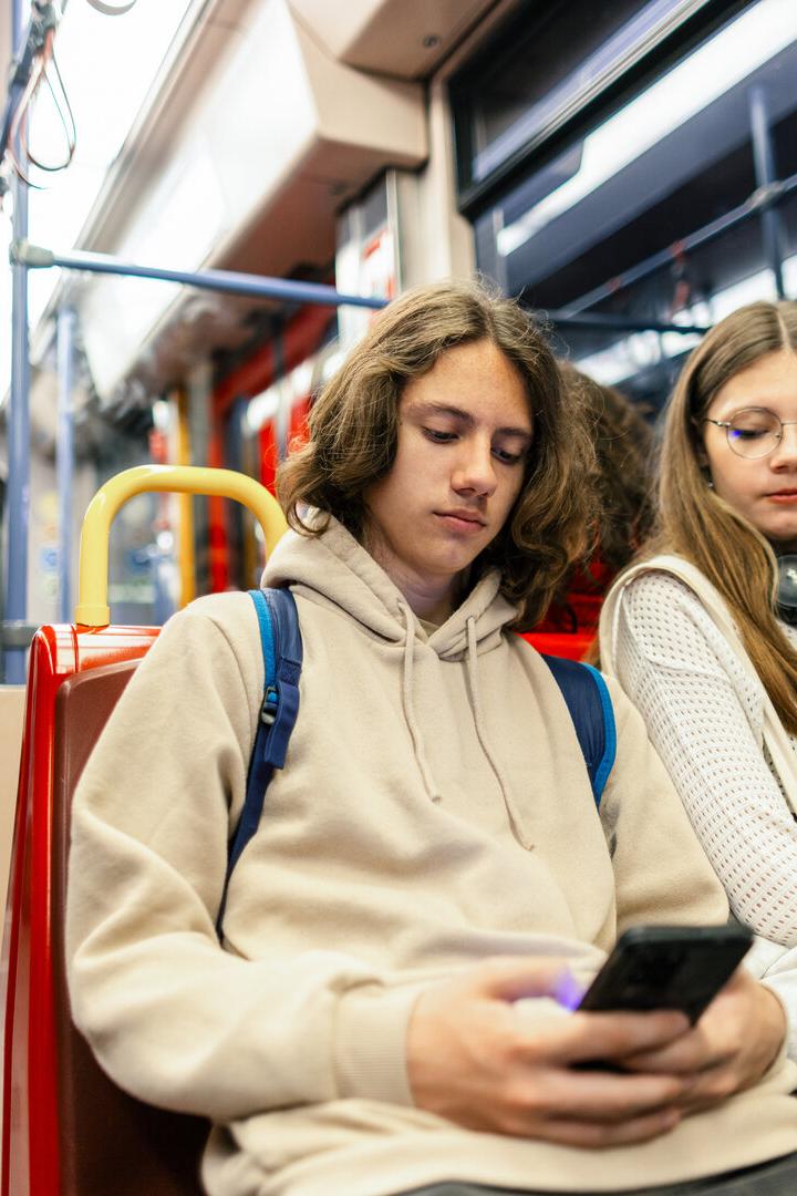 Zwei Jugendliche sitzen nebeneinander in einer Straßenbahn, einer schaut auf sein Handy, die andere blickt nach unten.