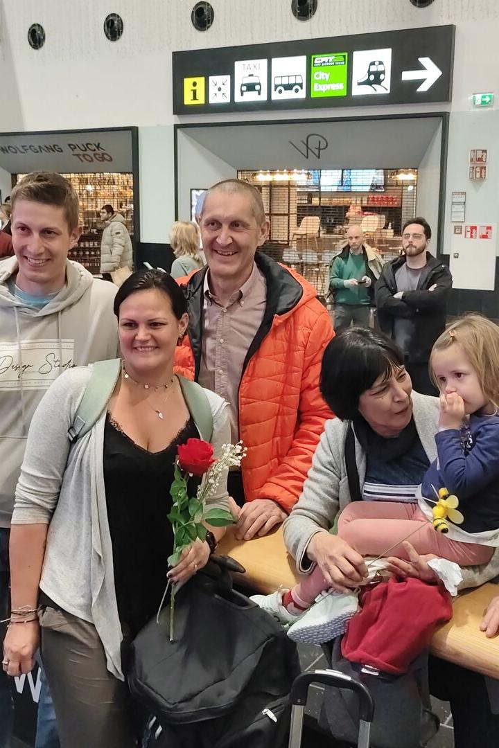 Sechs Menschen, darunter ein kleines Kind, stehen und sitzen lächelnd zusammen in einer belebten Bahnhofshalle.