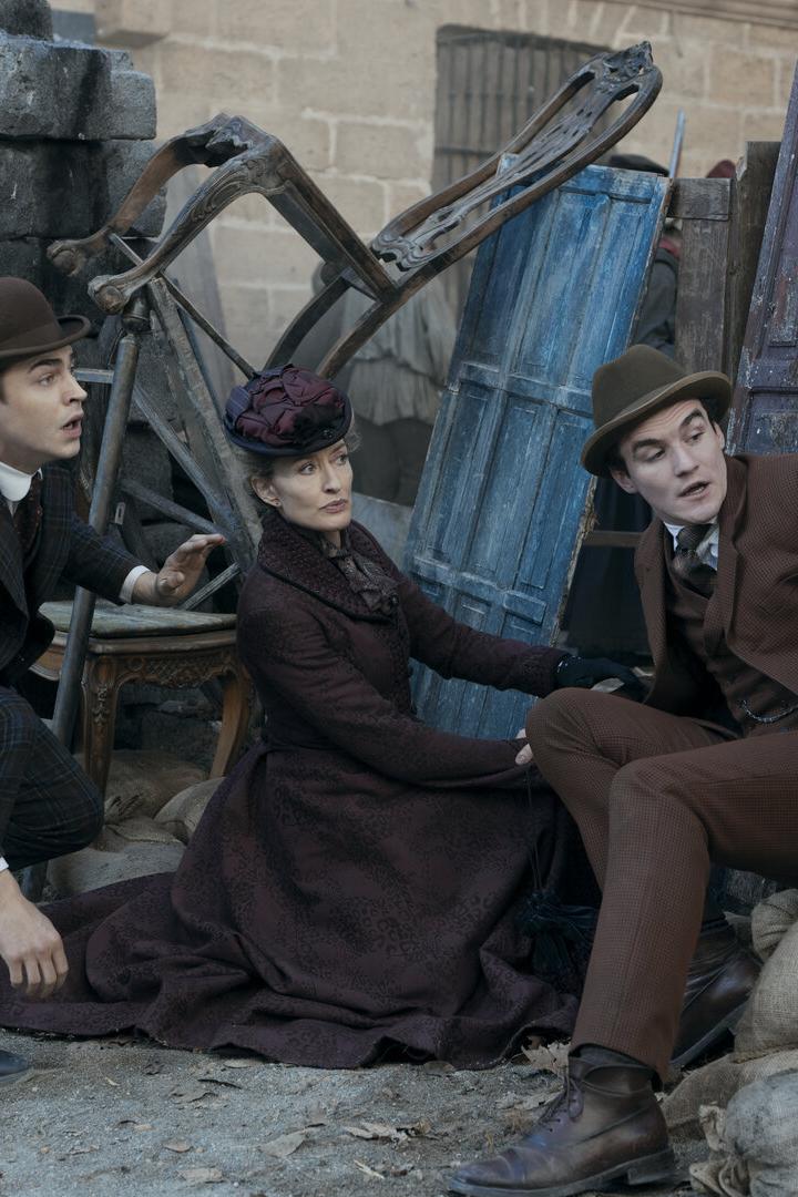 Drei Personen in historischer Kleidung ducken sich hinter Barrikaden aus Holz und Sandsäcken und wirken angespannt.