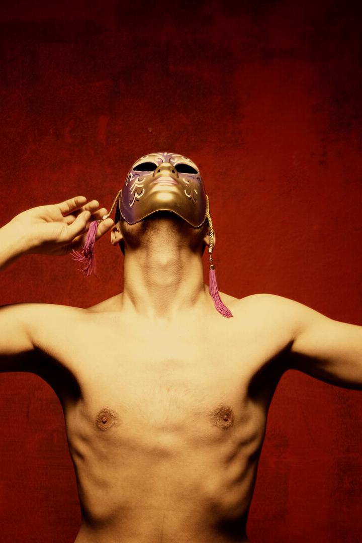 Oberkörper einer Person mit ausgebreiteten Armen und gold-violetter Maske vor rotem Hintergrund.