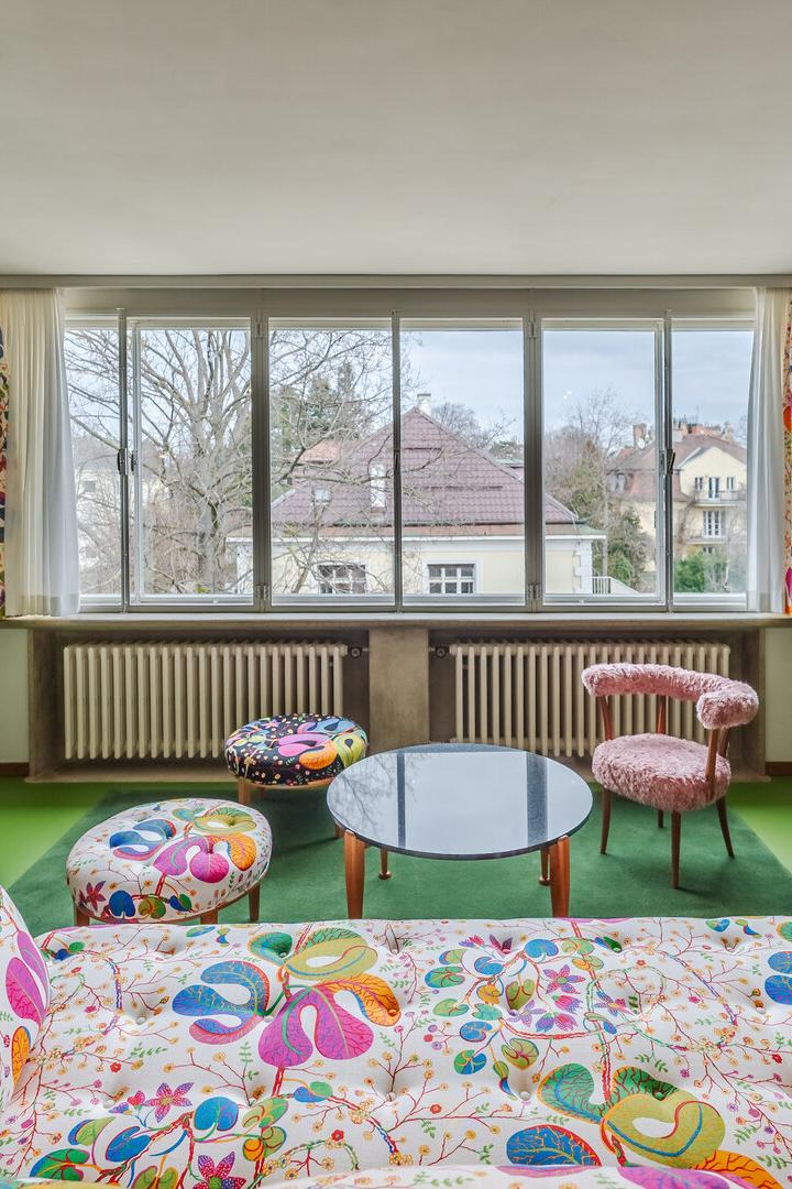 Wohnzimmer mit großen Fenstern, bunten Möbeln und Vorhängen, grünem Boden und Blick auf Häuser und Bäume draußen.