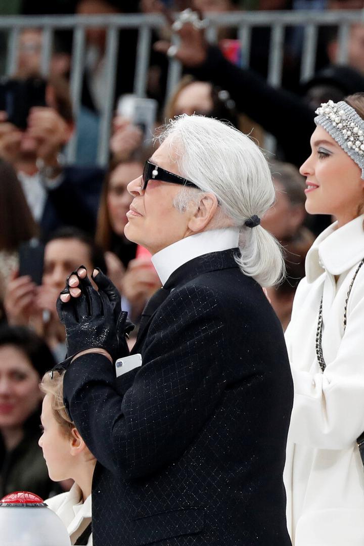 Karl Lagerfeld mit Sonnenbrille und Zopf, neben einem Model mit weißem Mantel, blickt aufmerksam in die Ferne.