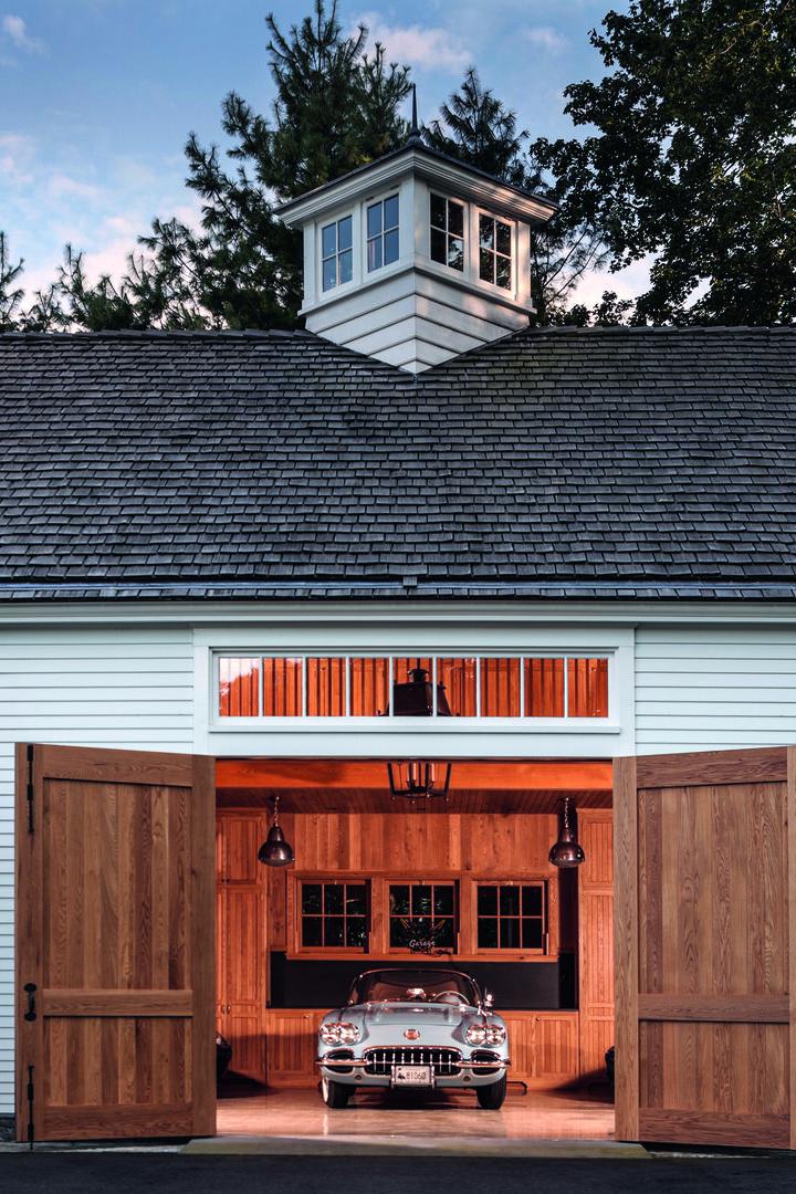 Ein Oldtimer steht in einer offenen, weiß gestrichenen Garage mit Holztoren und gepflegtem Außenbereich.