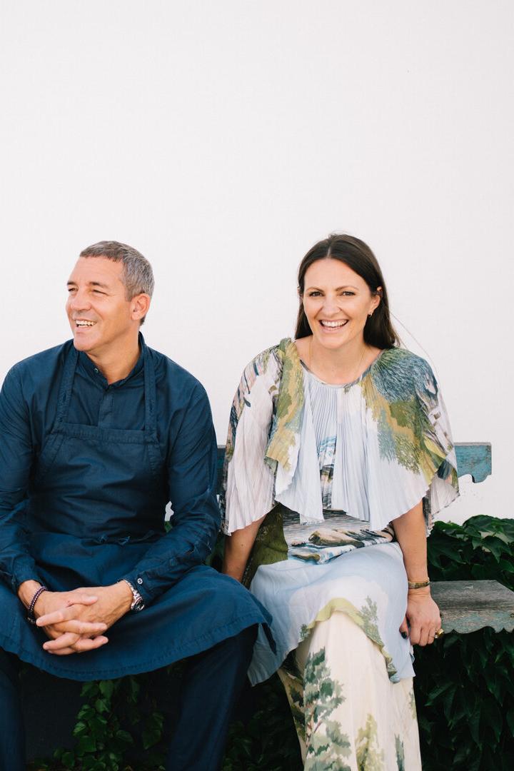 Ein Mann und eine Frau sitzen lachend auf einer alten Holzbank vor einer weißen Wand mit grünen Pflanzen im Hintergrund.